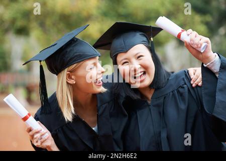 Wir haben endlich unseren Abschluss gemacht. Zwei Freunde in Togas draußen, die ihre Diplome hochhalten. Stockfoto