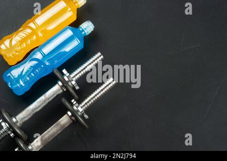 Plastikflaschen mit blauen und orangefarbenen isotonischen Getränken mit zwei Kurzhanteln auf dunklem Betonhintergrund, Kopierbereich. Stockfoto