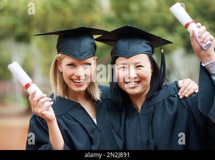 Wir haben endlich unseren Abschluss gemacht. Zwei junge College-Absolventen mit ihren Diplomen, während sie Mütze und Kleid tragen. Stockfoto