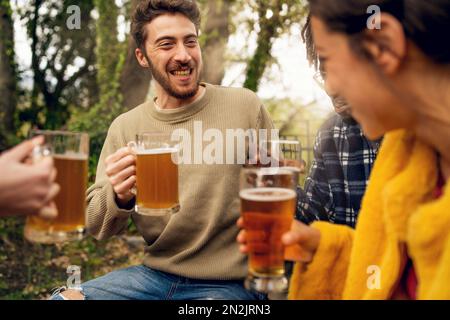 Eine Gruppe junger, multiethnischer Freunde entspannen sich bei einem Picknick und trinken Bier aus Steins. Der Fokus liegt auf einem Hipster mit Bart Stockfoto