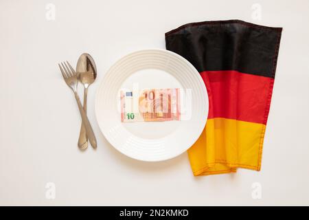 10 Euro liegen auf einem Teller neben einem Löffel und einer Gabel und der Flagge Deutschlands auf weißem Hintergrund, Finanzen und Wirtschaft in Deutschland, Wirtschaft Stockfoto