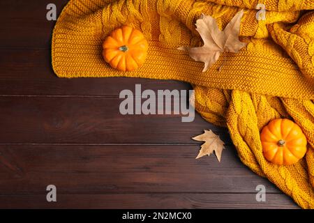 Orangefarbenes Strickkaro mit trockenen Blättern und Kürbissen auf braunem Holztisch, flach liegend. Platz für Text Stockfoto