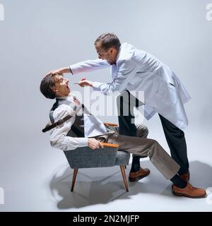 Junger Mann beim Zahnarzt, Doktor. Verrückter Arzt, der den Mund des Mannes überprüft. Modelle vor grauem Studiohintergrund. Ich habe Angst Stockfoto