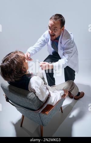 Doktor, Zahnarzt mit verrücktem Gesicht, der mit Zahnwerkzeugen in den Mund eines Mannes schaut, seine Zähne vor grauem Studiohintergrund überprüft Stockfoto