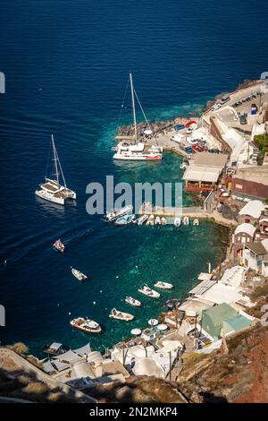 Oia, Griechenland - August 2022 : Vertikales Bild von Yachten, Booten und Katamaranen, die in einer kleinen Bucht in Oia auf der Insel Santorin vor Anker liegen Stockfoto