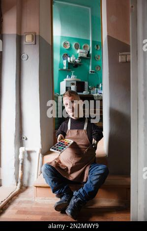 Porträt eines Mannes mit angeborener genetischer Erkrankung, der auf der Schwelle der Werkstatt mit einem Pinsel und Malfarben zum Zeichnen sitzt Stockfoto