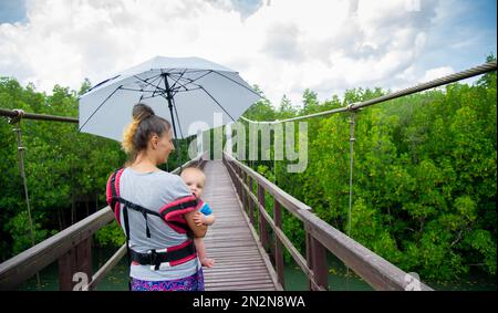 Touristen mit dazugehörigen Kindern gehen auf einer hölzernen Brücke durch Mangrovenwälder mit azurblauem Wasser im Chumphon National Park und schützen sich vor der Sonne Stockfoto