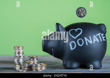 Seitenansicht der schwarzen Sparschweinbank mit dem geschriebenen Wort Spenden und signiertem Herz in der Nähe von Münzstapeln auf grauem Tisch auf grünem Hintergrund. Münzen fallen in Geld Stockfoto