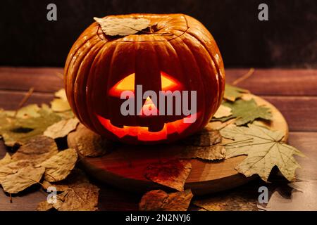 Halloween Kürbiskopf Jack Laterne auf dunklem Hintergrund mit trockenen Blättern. Leuchtende Gesichter sind ein Trick oder eine Belohnung Stockfoto