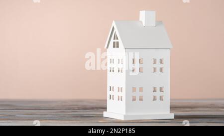 Weißes Spielzeugmodell eines Hauses aus Kartonpapier auf einem grauen Tisch auf pinkfarbenem Hintergrund. Immobilieninvestitionen. Wohnkosten, Versicherungszahlungen. Geschäftlich Stockfoto