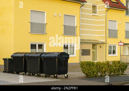 Schwarze Mülltonnen stehen in der Nähe eines gelben Wohngebäudes. Stockfoto
