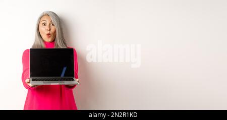 E-Commerce-Konzept. Beeindruckt asiatische ältere Frau zeigt leere Laptop-Bildschirm und starrte auf Kamera erstaunt, über weißem Hintergrund stehen Stockfoto