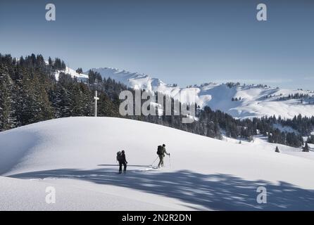 Zwei männliche Wanderer, die an einem übergroßen Kruzifix in Bergschnee vorbeilaufen, Ibergeregg, Schwyz, Schweiz Stockfoto