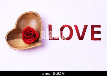 Rote Rose und das Wort Liebe aus rotem, glänzendem Papier auf dem Tisch. Grußkarte zum Valentinstag, zum Muttertag und zum Hochzeitstag Stockfoto