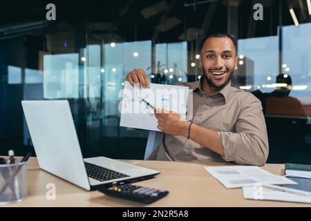 Portrait von jungen erfolgreichen Finanzierern, die im Büro arbeiten, lächelnd und vor die Kamera schauen, zeigt Grafik mit positiver Dynamik der wirtschaftlichen Gewinne, Geschäftsmann bei der Arbeit mit einem Notebook. Stockfoto
