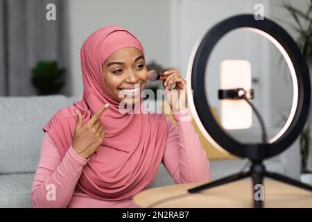Fröhliche schwarze muslimische Frau, die Kosmetik-Produktrezensionen aufnimmt, Puder aufträgt, Video für ihren Vlog per Telefon filmt Stockfoto