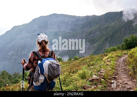 Rückansicht einer jungen Frau in kariertem Hemd und Mütze mit großem Rucksack mit Wanderstöcken, die im Sommer auf dem Bergpfad wandern und gesund und aktiv leben Stockfoto