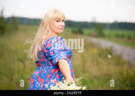 Schöne plus size Frau mit weißen Haaren in einem Sommerkleid posiert im Freien mit Gänseblümchen. Ein dickes Mädchen auf einer Wiese streut ihre Hand mit einem Strauß o aus Stockfoto