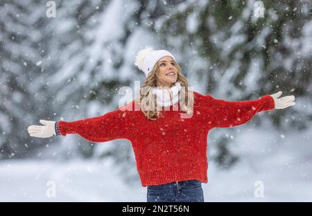 Mit weit geöffneten Armen genießt Frau in rotem Pullover, weißen Handschuhen, Schal und Hut an einem verschneiten Wintertag frische Luft. Stockfoto