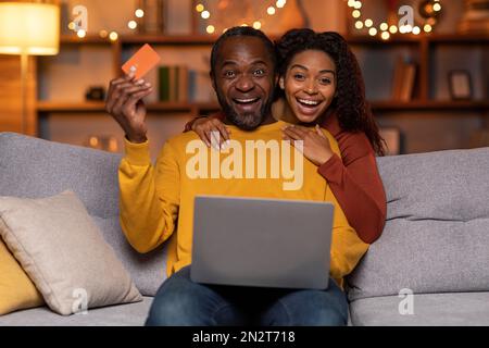 Aufgeregte afroamerikanische Ehepartner, die ihren Laptop benutzen und ihre Kreditkarte vorzeigen Stockfoto