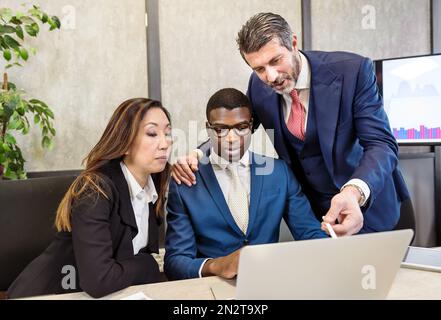 Konzentrierte Mitarbeiter verschiedener Rassen in formeller Kleidung, die an Geschäftsstrategien arbeiten, während sie während der Zusammenarbeit in modernen Arbeitsbereichen ein Notebook verwenden Stockfoto