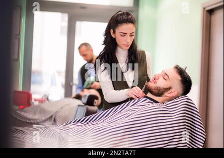 Konzentrierte professionelle Friseurin, die den Bart mit der Schere des männlichen Klienten in einem modernen Friseursalon justiert Stockfoto