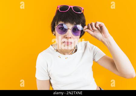 Tarnkonzept. Ein junges Mädchen mit mehreren Sonnenbrillen, das verdächtig in die Kamera schaut und eine Sonnenbrille in der Hand hält. Studioaufnahme. Orangefarbener Hintergrund. . Hochwertiges Foto Stockfoto
