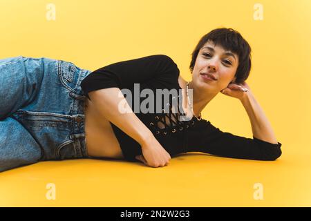Selbstbewusste, kurzhaarige Frau, die auf dem Boden liegt und in die Kamera schaut. Hochformat mit niedrigem Winkel. Studio-Aufnahmen vor gelbem Hintergrund. Hochwertiges Foto Stockfoto