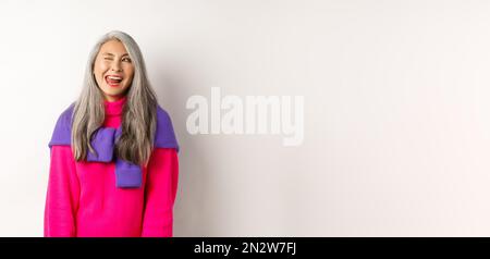 Lustige und glückliche asiatische ältere Frau mit Spaß, zeigt Zunge und winken frech, Blick links auf Logo, stehend über weißem Hintergrund Stockfoto
