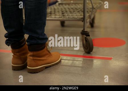Person mit Warenkorb, die im Store hinter einer Bodenmarkierung mit Klebeband steht, um soziale Distanz zu wahren, Nahaufnahme. Präventive Massnahmen während der Coronavirus-Pandemie Stockfoto