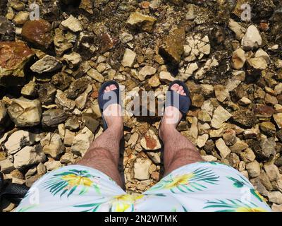 Lustiges Sommerfoto von Füßen in blauen Flip-Flops und bunten Badehosen Stockfoto