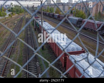 Sehen Sie durch den Netzzaun über einen langen Güterzug mit Wagen ohne Dächer, beladen mit riesigen weißen Säcken und warten am Bahnhof in Ky Stockfoto