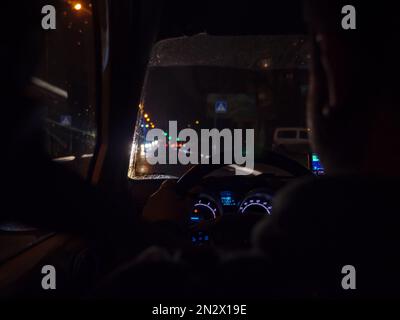 Hervorgehobenes Armaturenbrett eines Autos, das durch dunkle Nachtstädte fährt, mit verschwommenem Fahrer hinter dem Steuer, Regentropfen auf einer Windschutzscheibe, Lichter im Straßenverkehr. Stockfoto