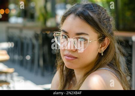 Porträt einer jungen venezolanischen Frau mit Brille, gelangweilt, müde, verärgert und ernst, die in die Kamera schaut und auf das Essen im Restaurant wartet. Stockfoto