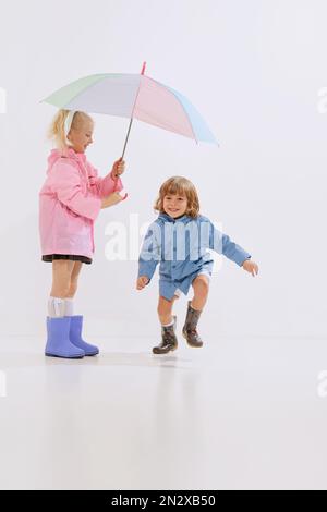 Süße Kinder, glückliche Kinder in Regenmänteln spielen, Spaß zusammen haben isoliert auf weißem Hintergrund. Konzept von Kindheit, Freundschaft, Familie, Spaß Stockfoto