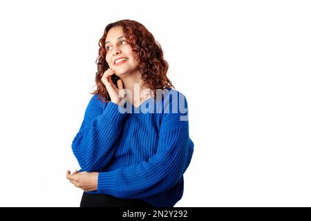 Ein junges Mädchen, das glücklich lächelt, einen blauen Pullover trägt, isoliert auf weißem Hintergrund, wegsieht, lächelt, selbstbewusst aussieht. Berührt das Kinn. Konzept Stockfoto