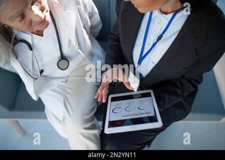 Aus einem großen Blickwinkel betrachtet, wie eine Geschäftsfrau dem Arzt ein Diagramm auf einem digitalen Tablet zeigt. Stockfoto