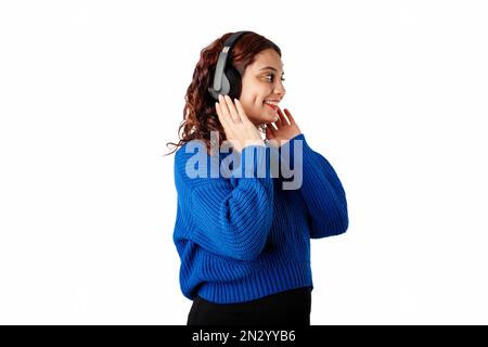 Seitenansicht einer jungen Frau mit blauem Pullover, isoliert auf weißem Hintergrund und berührendem Headset, mit Blick auf den leeren Kopierbereich und sehr zufrieden. Stockfoto