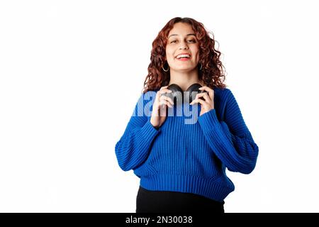 Junge weiße Frau mit Pullover-Pullover, die isoliert auf weißem Hintergrund steht und das Headset um den Hals hält, in die Kamera schaut und lacht Stockfoto