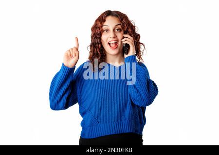 Fröhliche Frau, die einen blauen Pullover trägt, isoliert auf weißem Hintergrund und eine Idee oder Frage hat, während sie mit dem Finger nach oben auf das Mobiltelefon zeigt. Erhält V Stockfoto