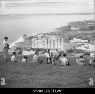 1955, historisch, ein männlicher Lehrer mit einer kleinen Gruppe von Jugendlichen Schülern auf einem Ausflug auf dem grünen Hügel mit Blick auf Alum Bay auf der Isle of Wight, Hampshire, England, Großbritannien. Stockfoto