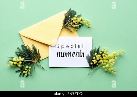 Text Special Moments auf eine Papierkarte mit Mimosablüten in einem gelben Umschlag auf minzgrünem Hintergrund. Natürliche Einrichtung im Frühling. Flach liegend, Draufsicht. Stockfoto