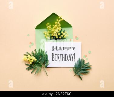 Text Happy Birthday (alles gute zum Geburtstag) auf einer Papierkarte, Mimosablüten in grünem Umschlag auf gelbem goldenen Hintergrund. Flach liegend, zentrale Zusammensetzung. Stockfoto