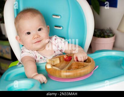 Baby isst Brokkoli. Säuglingsprothetik. Entwöhnung. Gesunde Ernährung. Kaukasisches Mädchen, das in einem Hochstuhl sitzt und ihr Mittagessen isst Stockfoto