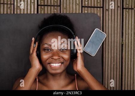 Porträt einer jungen, wunderschönen, lächelnden afrikanerin mit Kopfhörern, die in die Kamera schaut, während sie auf einer Yoga-Matte liegt Stockfoto