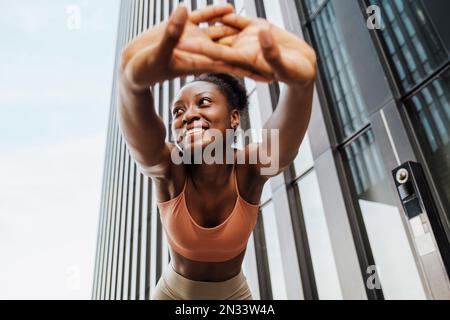 Junge, schöne, sportliche, lächelnde, glückliche afrikanerin, die sich mit verflochtenen Händen den Rücken streckt und zur Seite schaut, während sie draußen steht Stockfoto