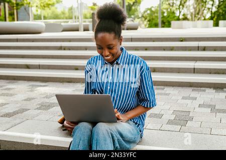 Junge, wunderschöne, lächelnde afrikanerin, die mit einem Laptop arbeitet, während sie draußen auf einer Treppe sitzt Stockfoto