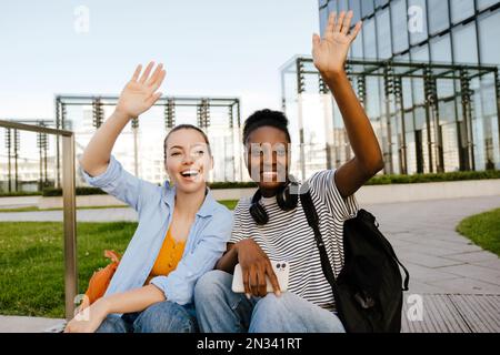 Junge, multiethnische Frauen, die lächeln und sich die Hände winken, während sie auf der Straße der Stadt sitzen Stockfoto