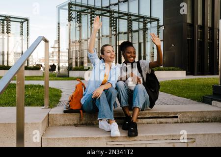 Junge, multiethnische Frauen, die lächeln und sich die Hände winken, während sie auf der Straße der Stadt sitzen Stockfoto