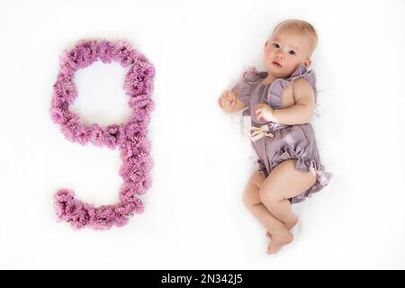 Kleines neugeborenes Mädchen in einem lila Strampelanzug. 9 Monate altes Baby. Neun Monate Baby-Meilenstein. Stockfoto